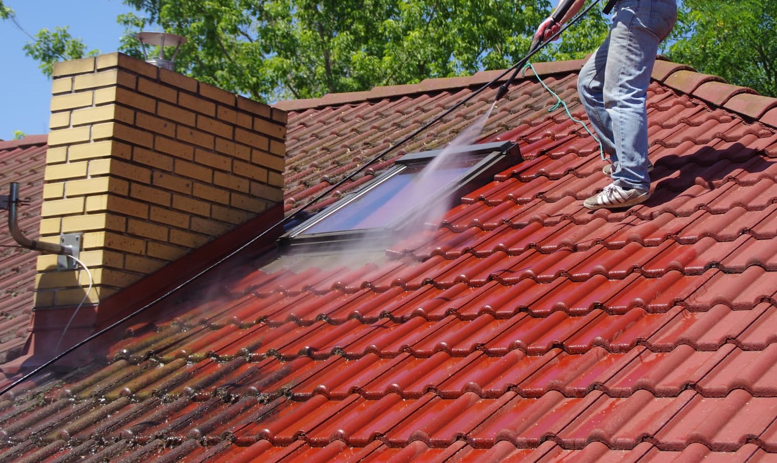 Pressure-washing moss off red roof tiles beside a chimney, roof cleaning and moss removal